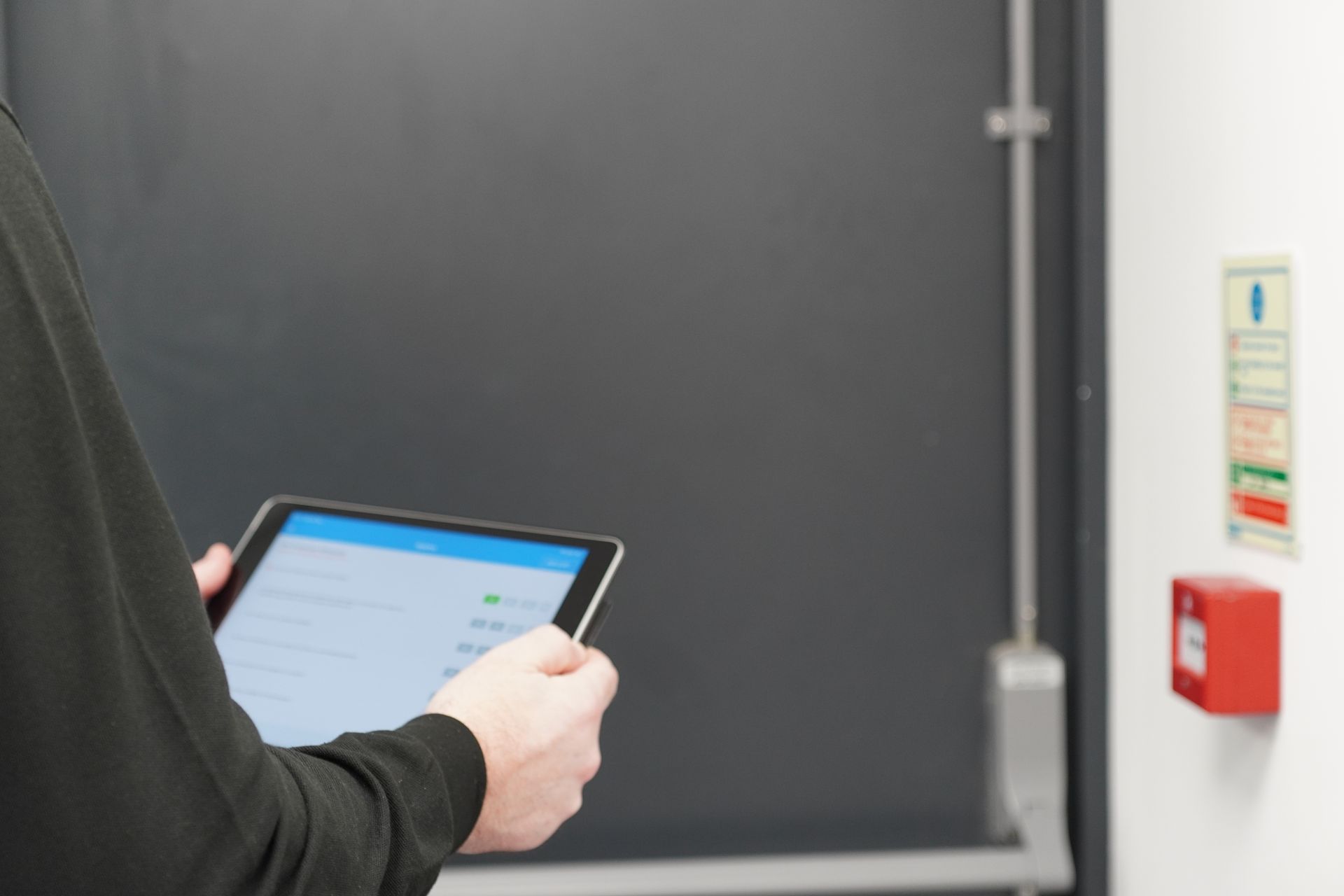 Person holding a tablet, inspecting a door, with fire safety equipment visible.