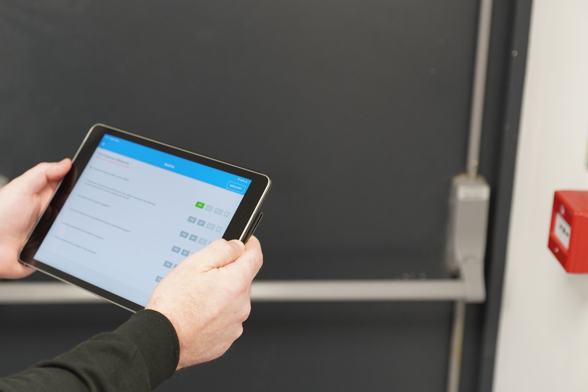 Person holding a tablet, viewing a screen with data, near a door and fire alarm.