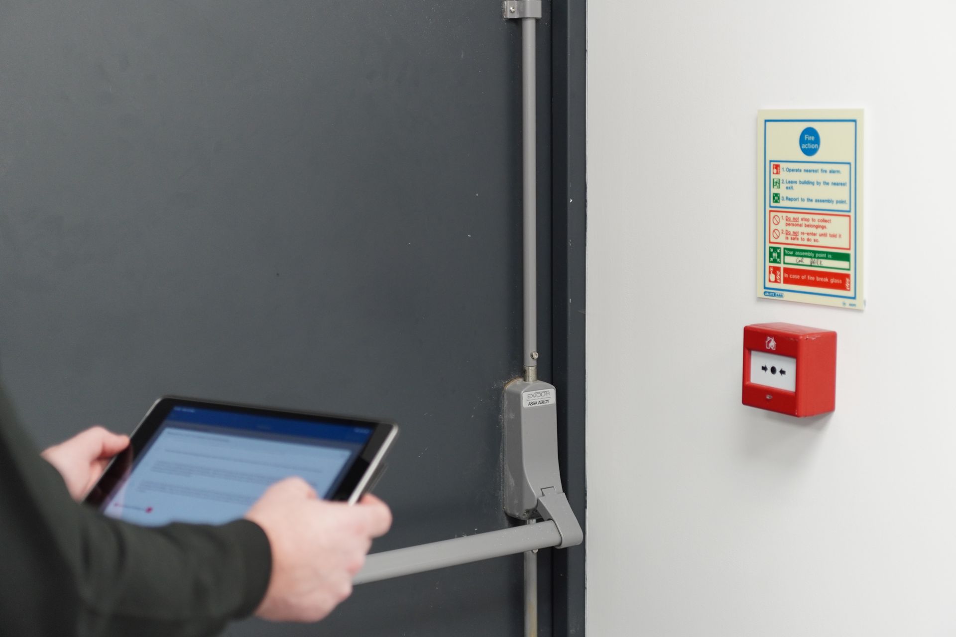 Person using tablet near a fire exit door, fire alarm, and safety instructions on a white wall.