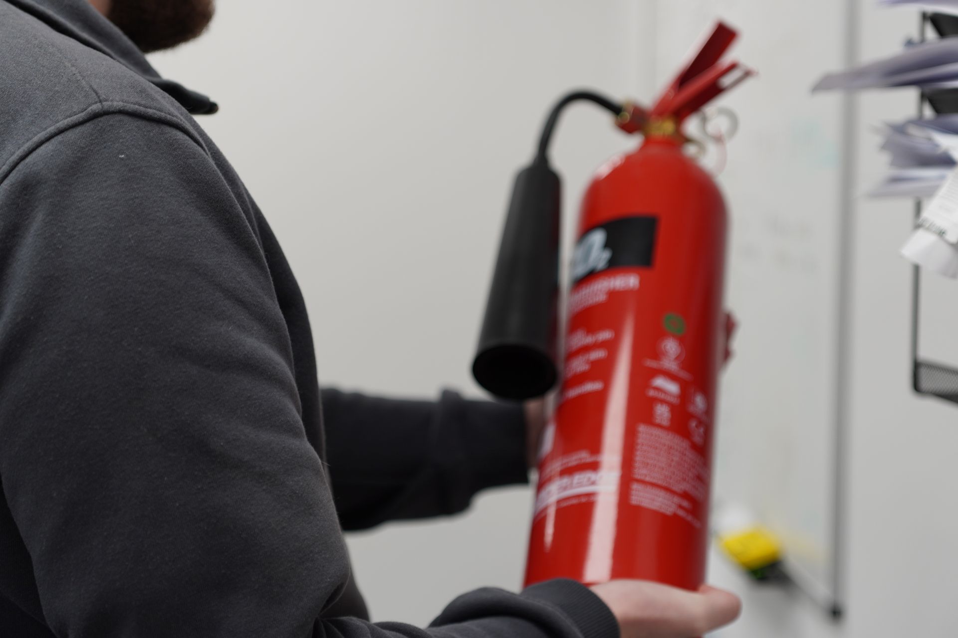 Person holding a red fire extinguisher, black nozzle, in a white-walled office setting.