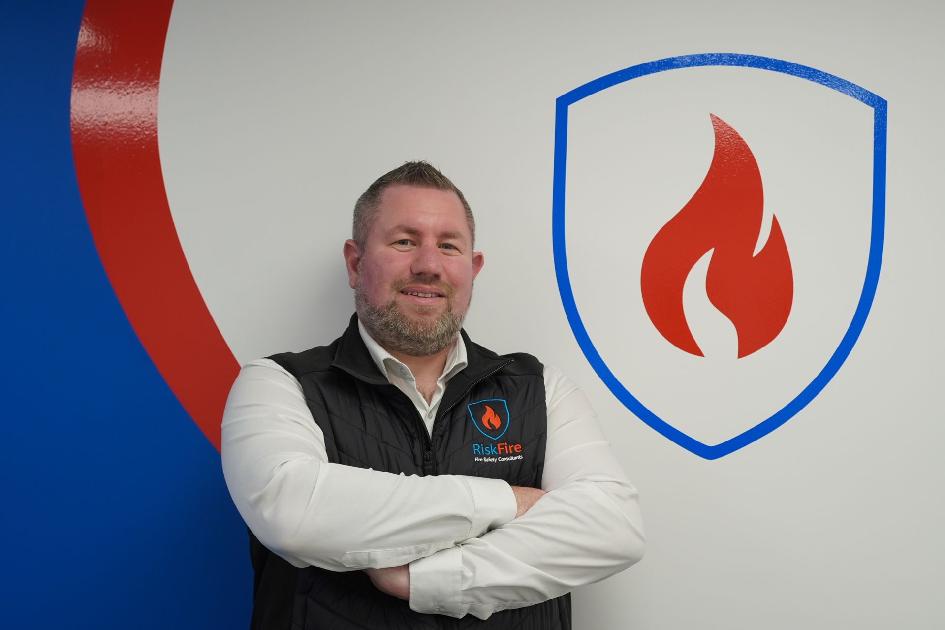 Man in vest with arms crossed, standing in front of a company logo with a flame and shield, indoors.