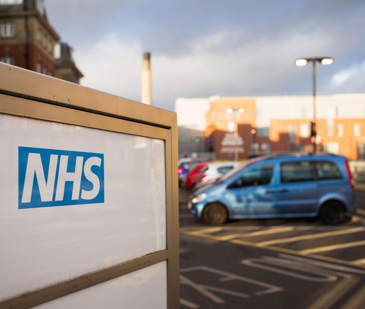 NHS sign with blurred hospital and cars in the background.
