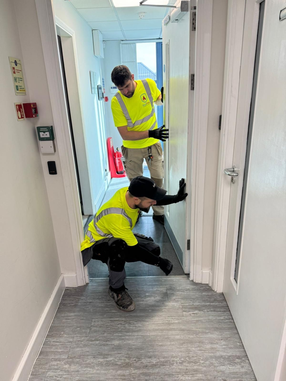 Two workers in neon vests install a door in a hallway. One crouches, the other stands, both using hands.