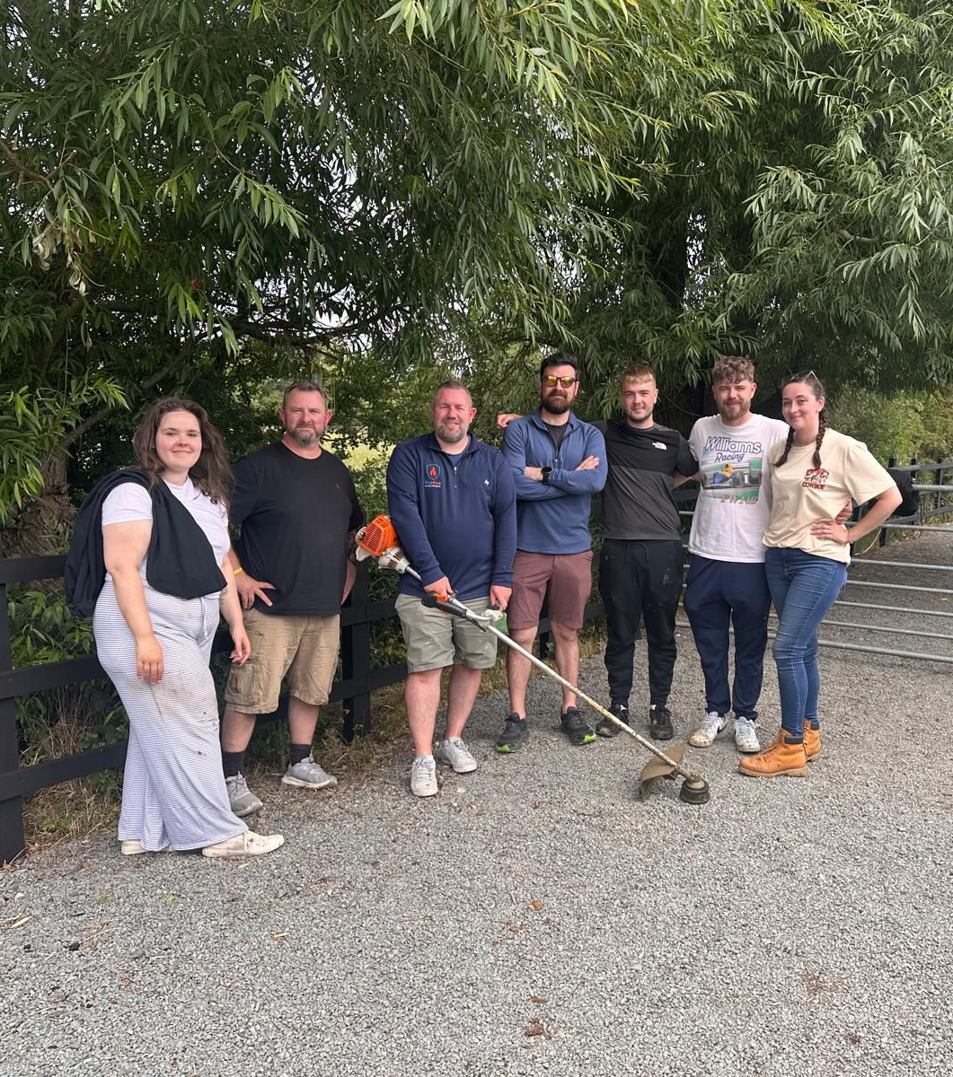 Group of eight people outdoors with a weed whacker, smiling, near trees and a fence.