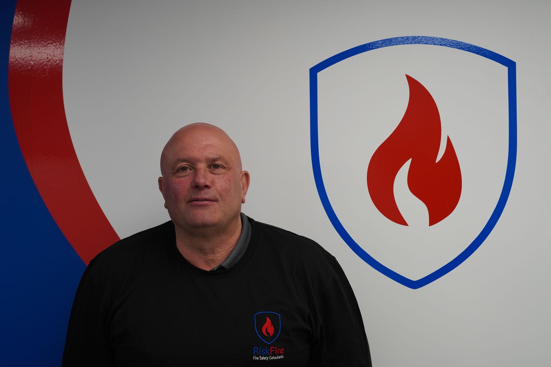 Man in black polo shirt with crossed arms, smiling, in front of a blue and red shield graphic.