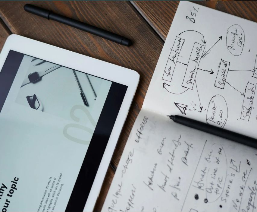 Tablet and notepad with handwritten notes and diagrams, next to two pens on wood.