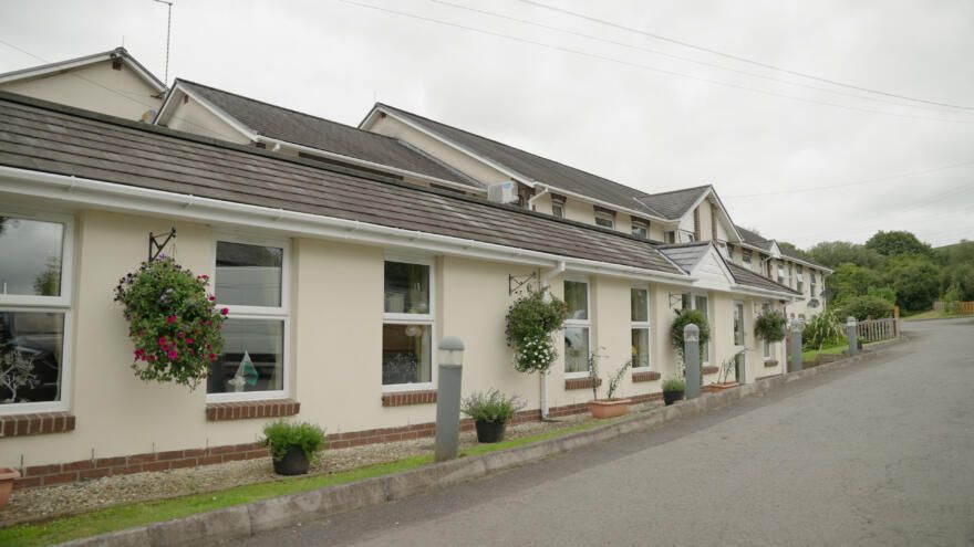 Cream-colored building with dark roof tiles, windows, hanging flower baskets, and a road in front.