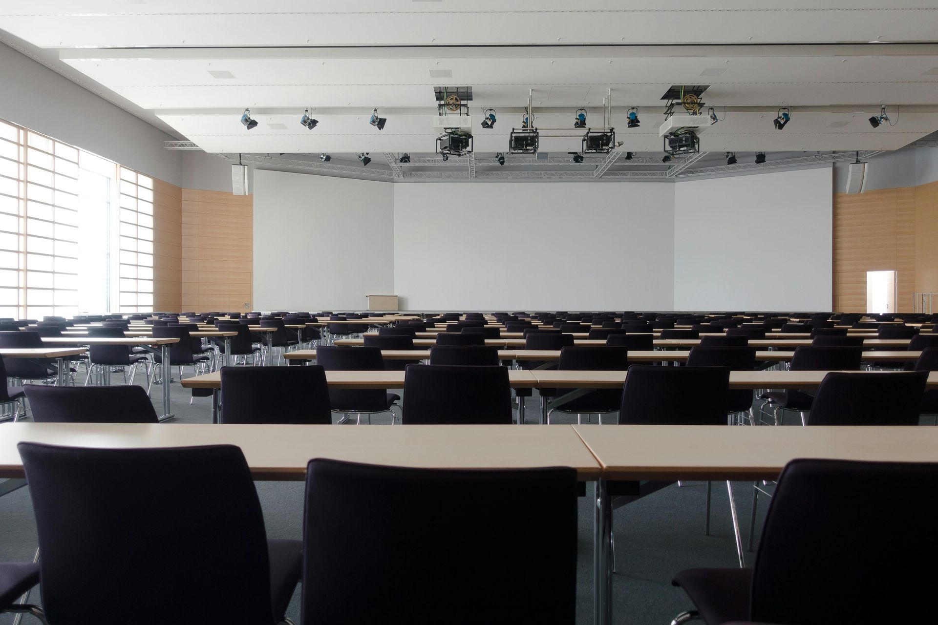 Empty auditorium with rows of tables and chairs facing a large screen and stage.