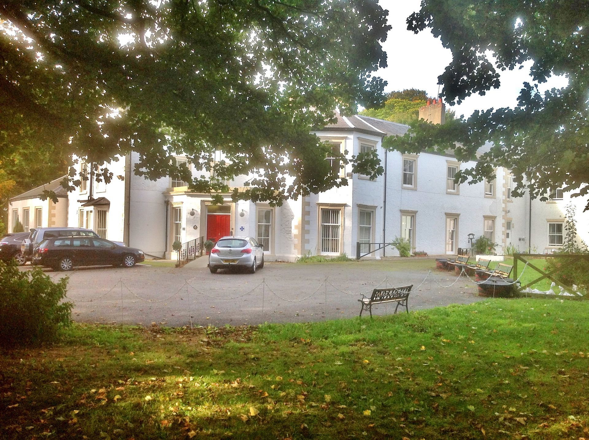 White two-story building with red front door, cars in a circular drive, under a tree on a grassy lawn.