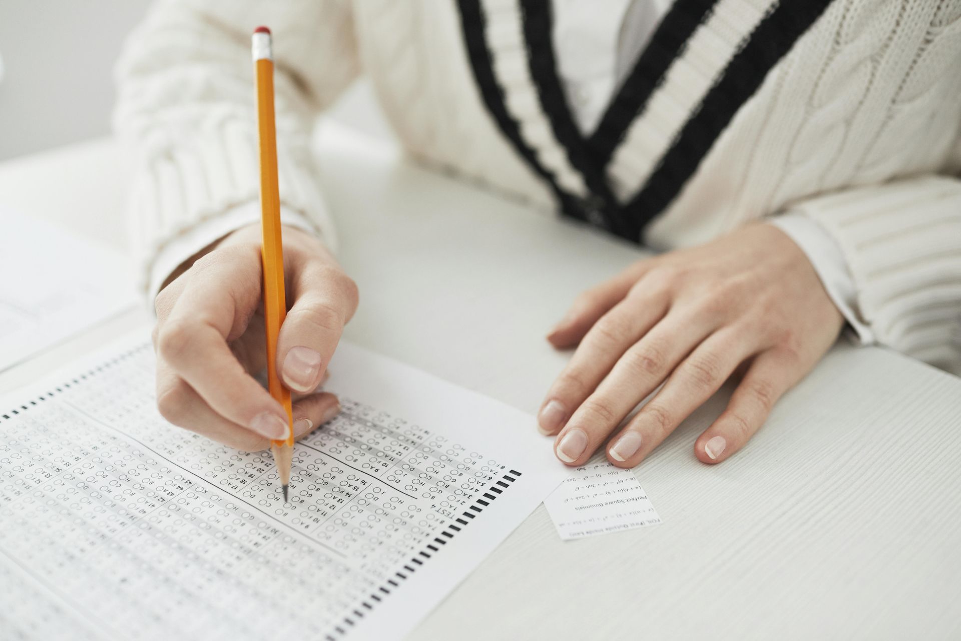 Person writing on a scantron test with a pencil, wearing a white sweater.