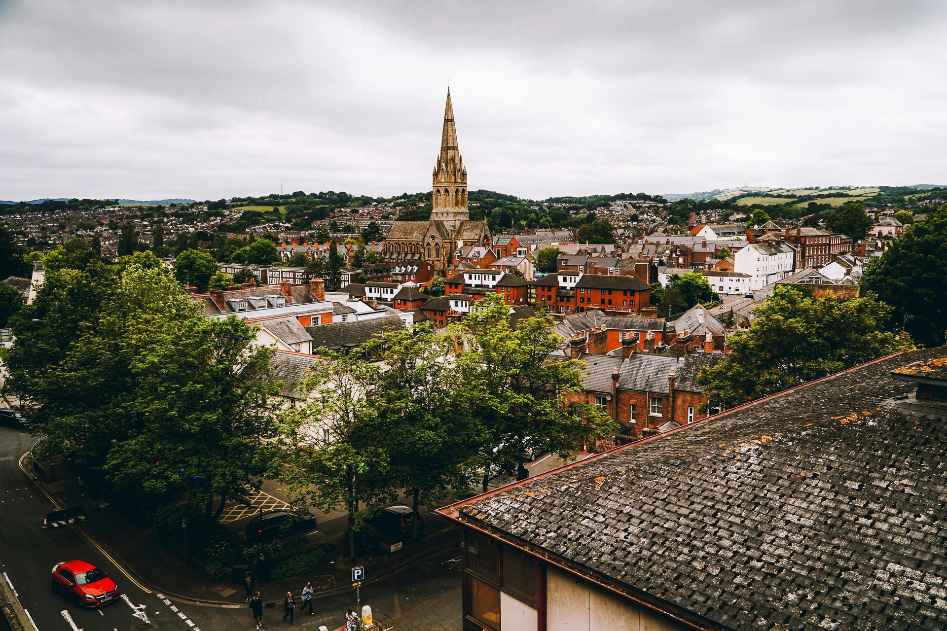 View of a town with a tall church spire, red-roofed buildings, green trees, and a cloudy sky.