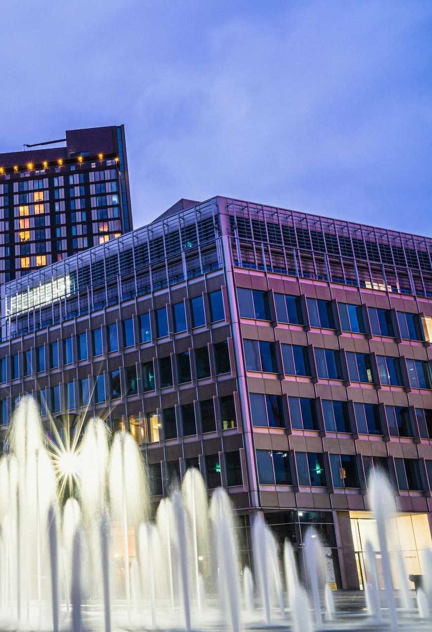Fountain sprays in front of office buildings at dusk, under a blue sky.