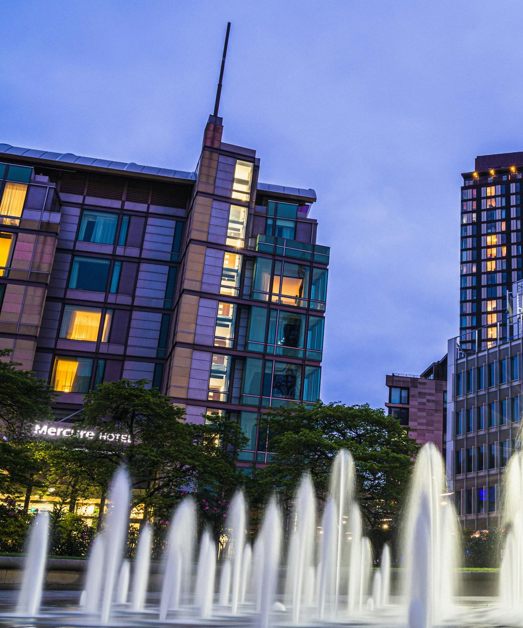 Fountain sprays in front of modern buildings at dusk. Teal glass facade reflects warm lights, blue sky overhead.