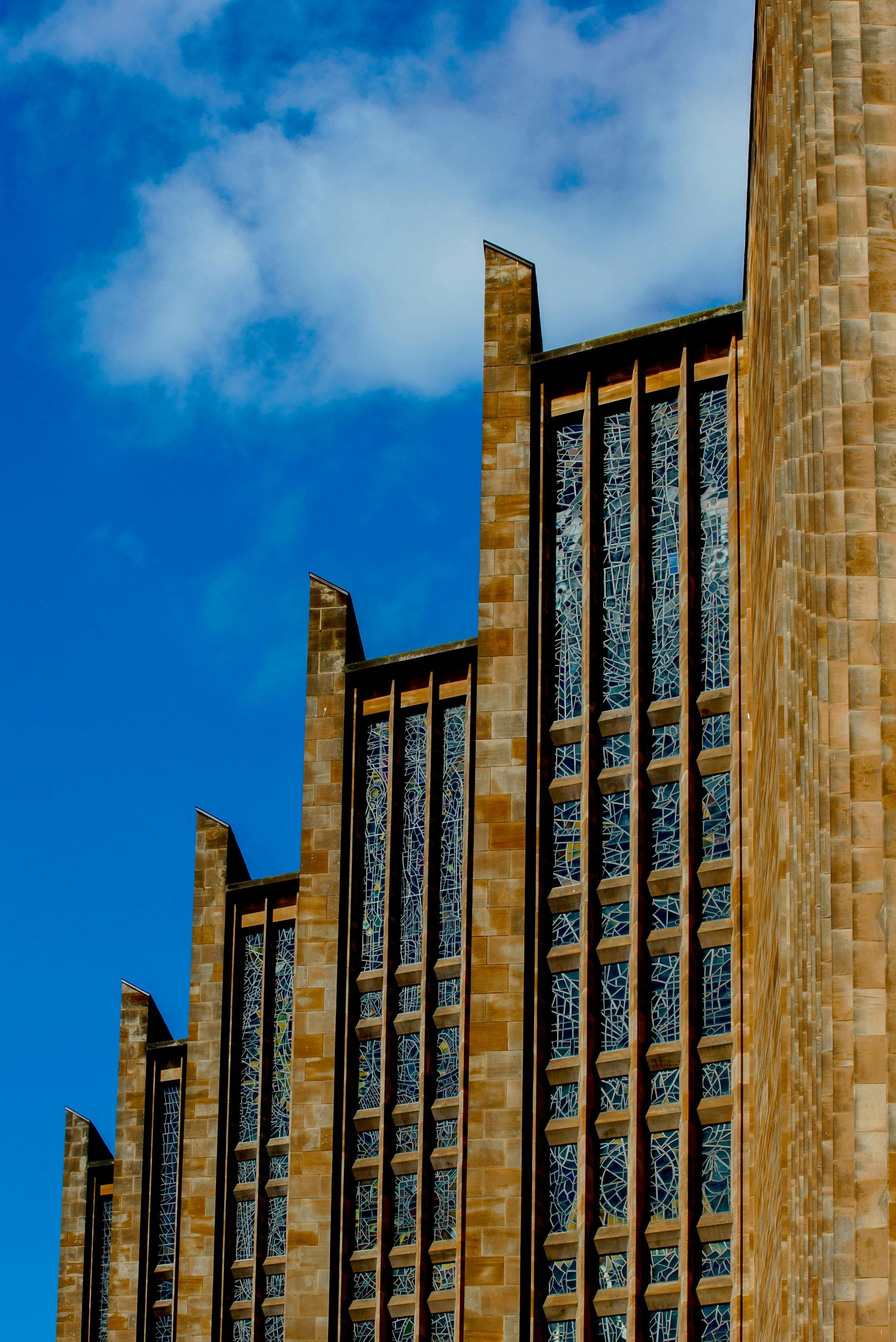 Tall sandstone building with many tall, narrow windows against a bright blue sky with clouds.