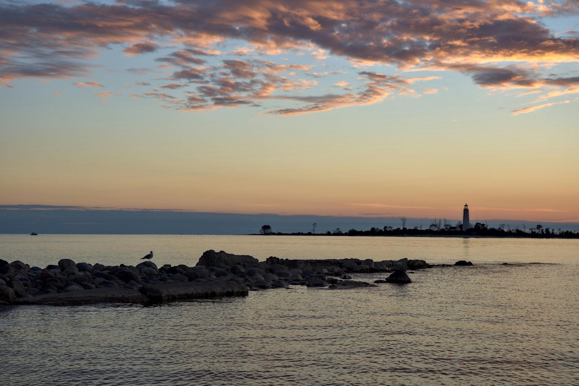 Sunset over calm water with rocky shoreline and distant skyline. Sky features orange and pink clouds.