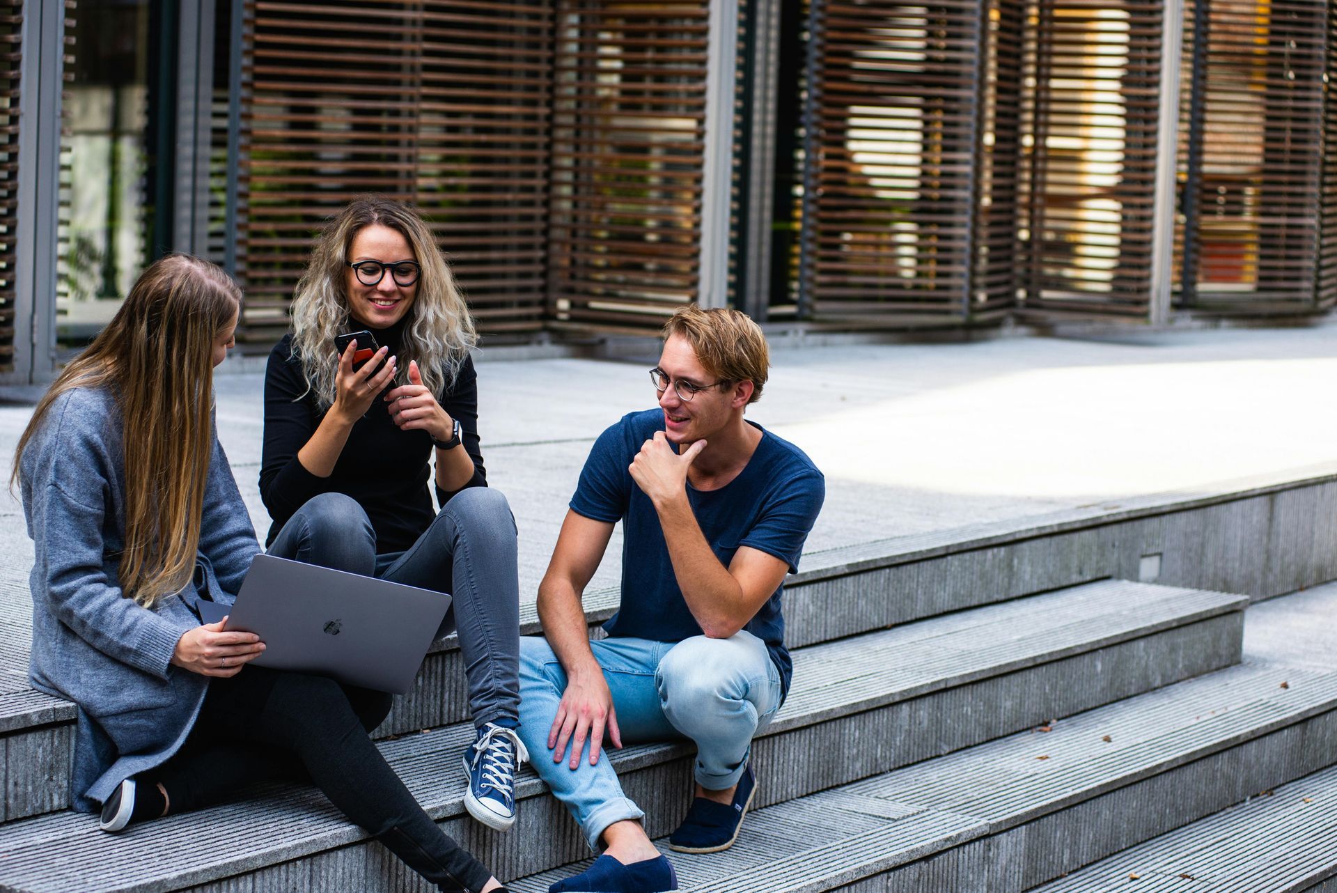Three people sitting on outdoor steps, one using a laptop, one on a phone, and one smiling and talking.