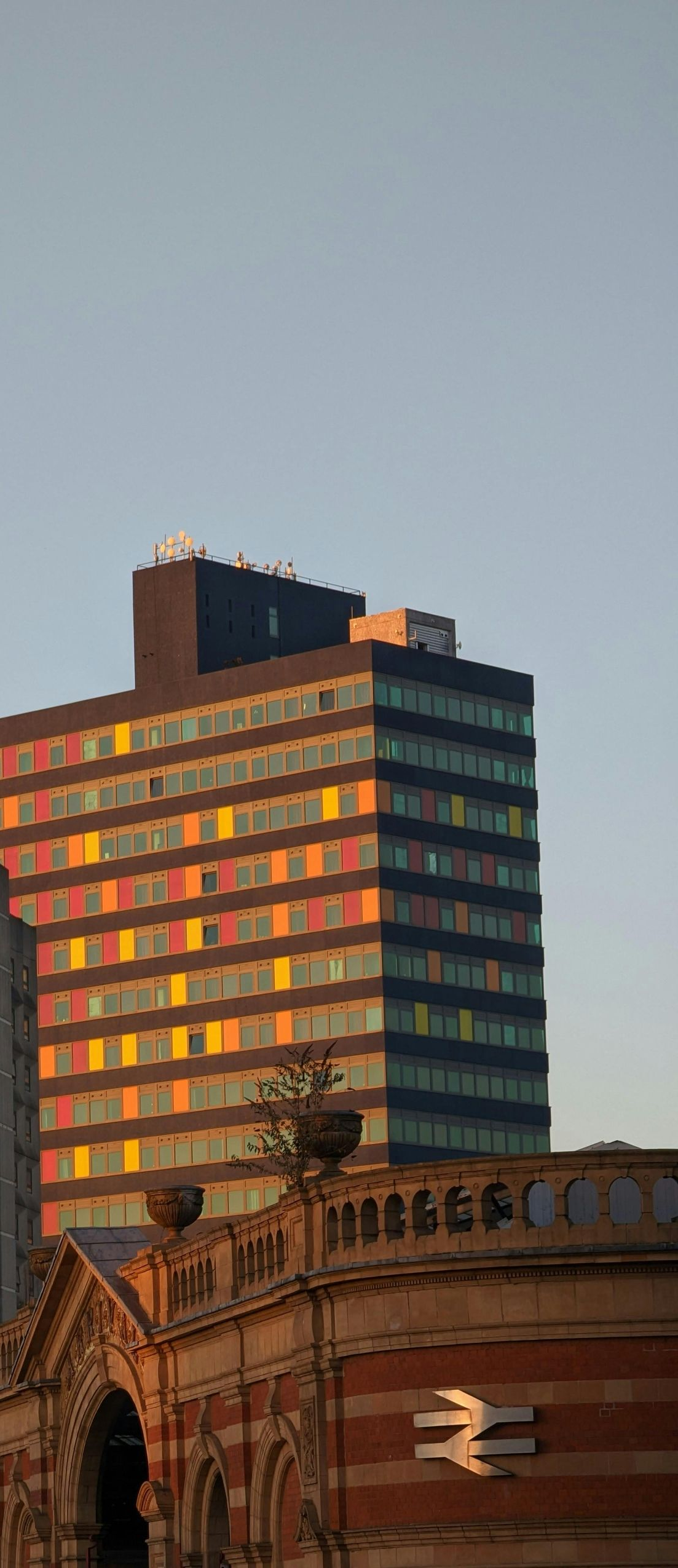 A tall building with colorful windows behind a building with brickwork, under a light blue sky.