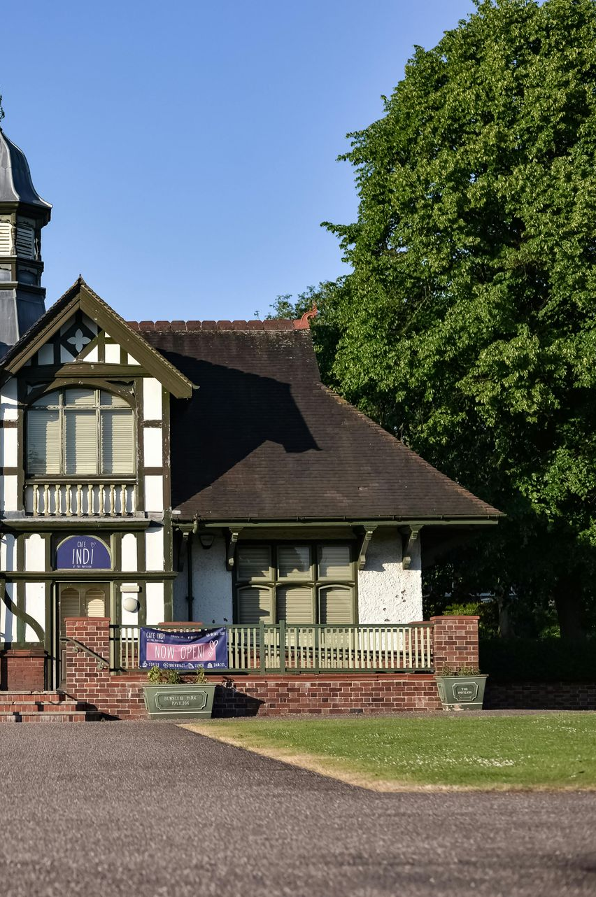 Tudor-style building with white and dark wood trim, brick base, and green accents next to a lush tree on a sunny day.