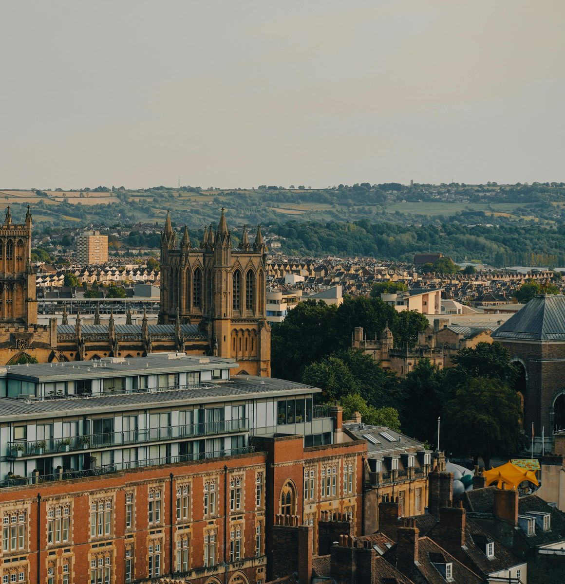 Cityscape with church towers, buildings, and green hills in the background under a light sky.