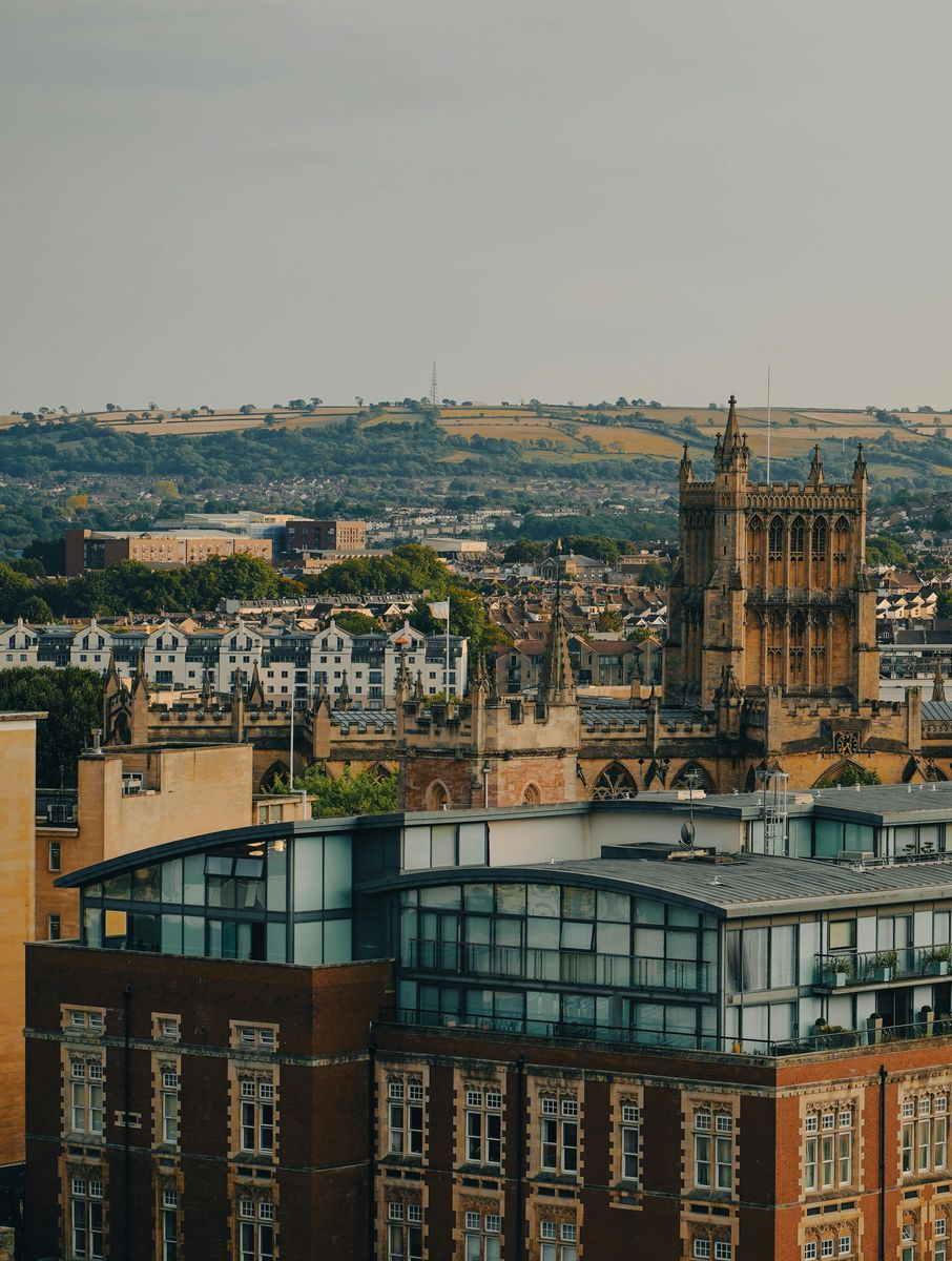Cityscape with a tall church tower, buildings, and a distant hilly landscape under a cloudy sky.