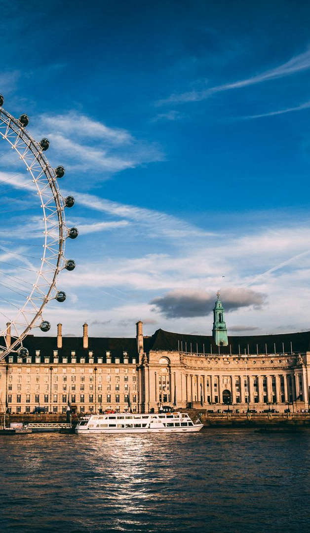London Eye with building and boat along the Thames River under blue sky.