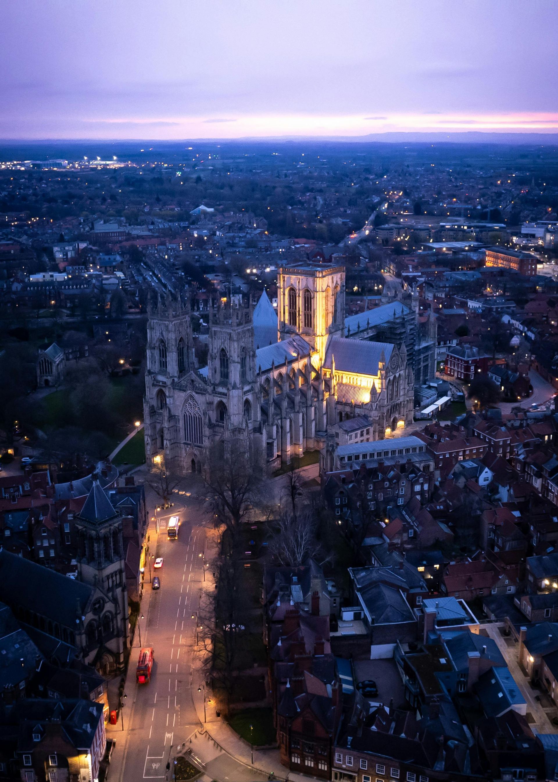York Minster at dusk, illuminated against a dusky purple sky, surrounded by city buildings and streets.