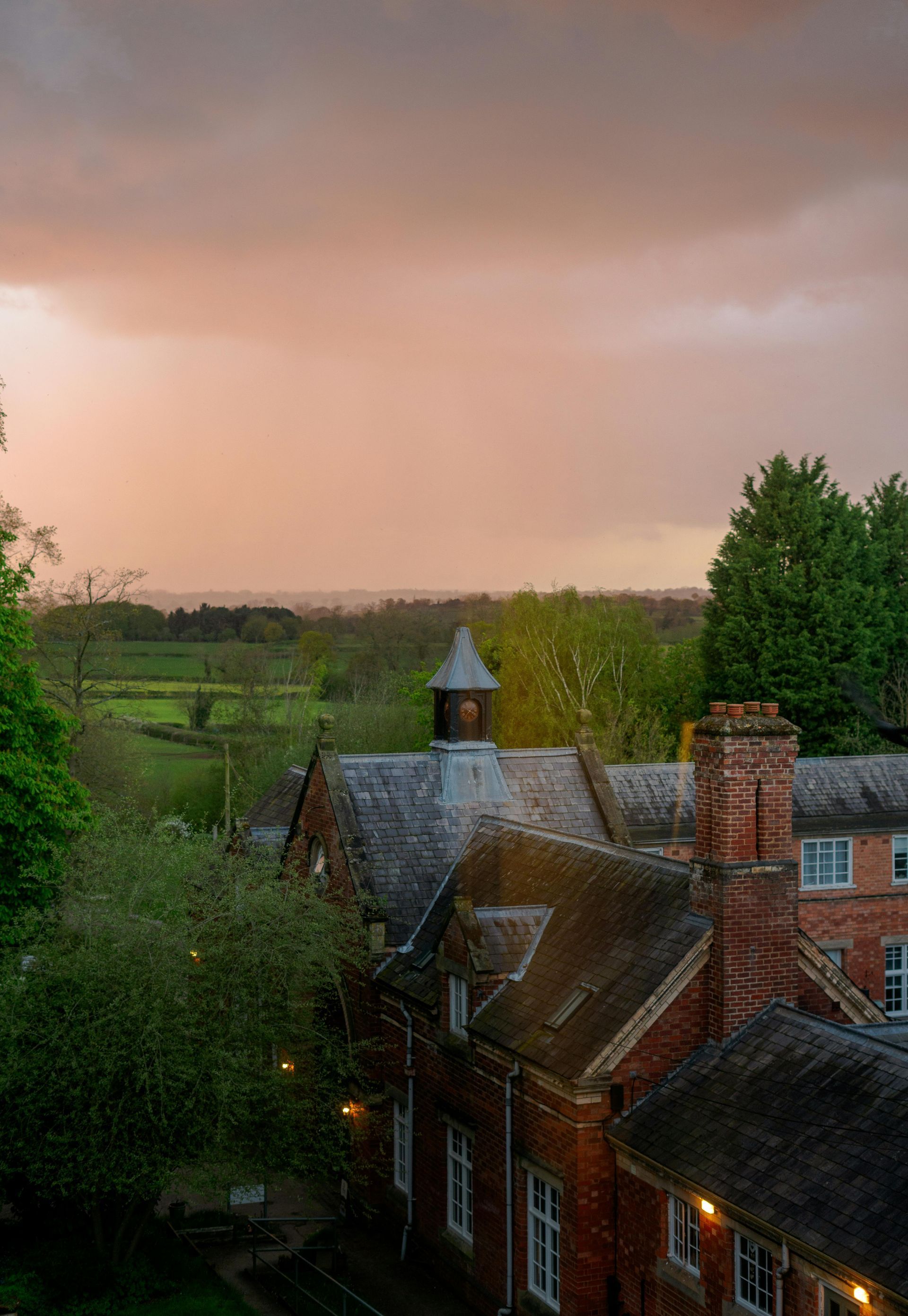 Brick building roofs and chimney with green trees and fields under an orange and gray sky.
