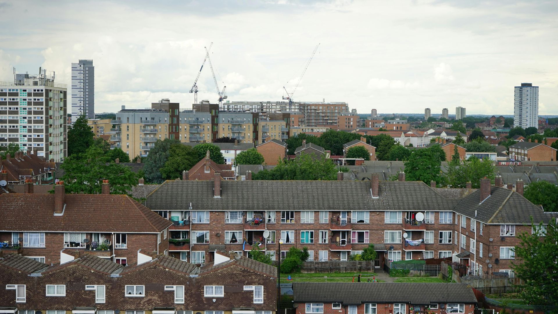 Cityscape with low-rise housing in foreground, taller buildings and construction cranes in the background under a cloudy sky.