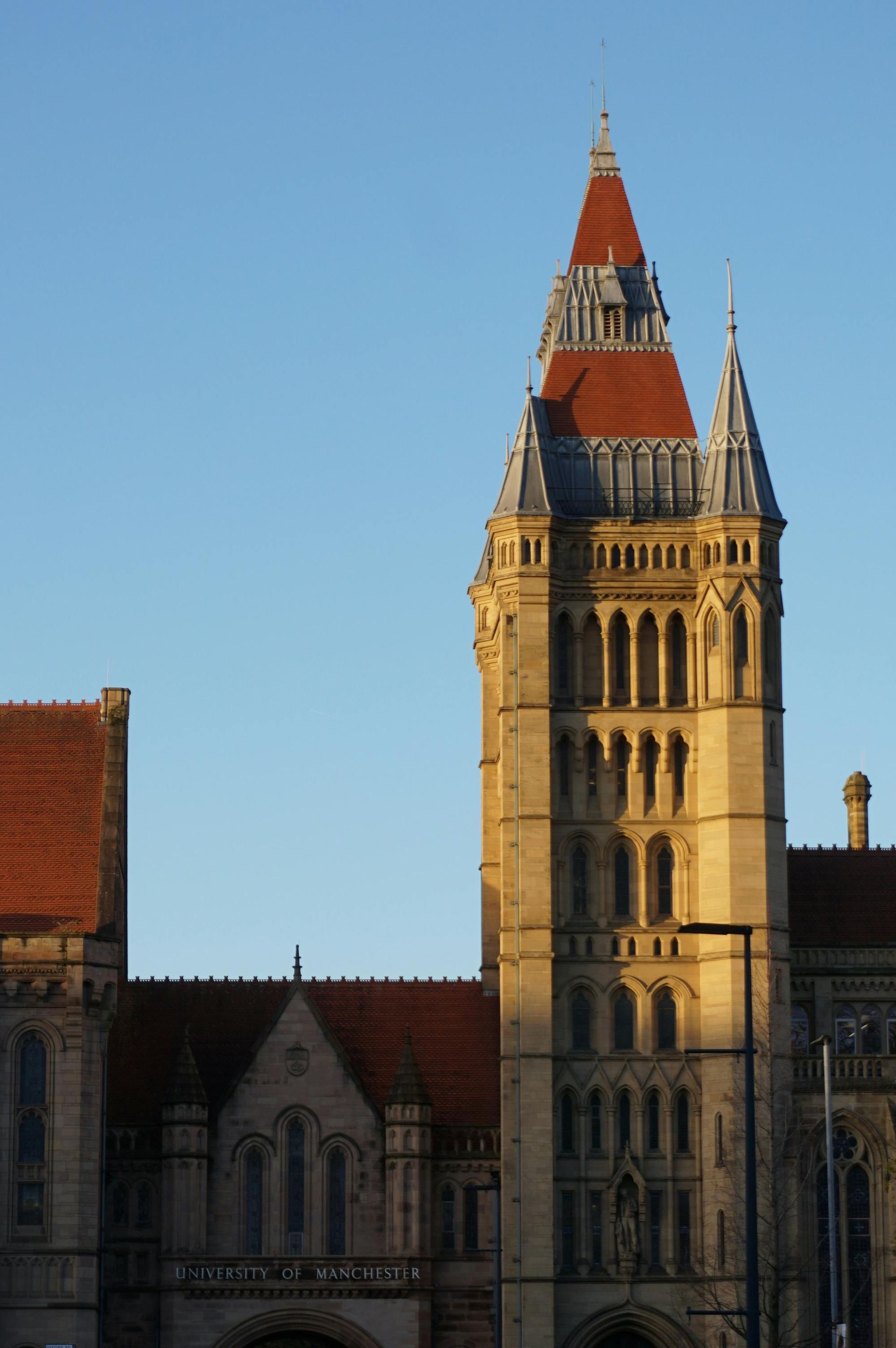 Tall tower with a red-tiled roof, part of a stone building, illuminated by sunlight against a clear blue sky.
