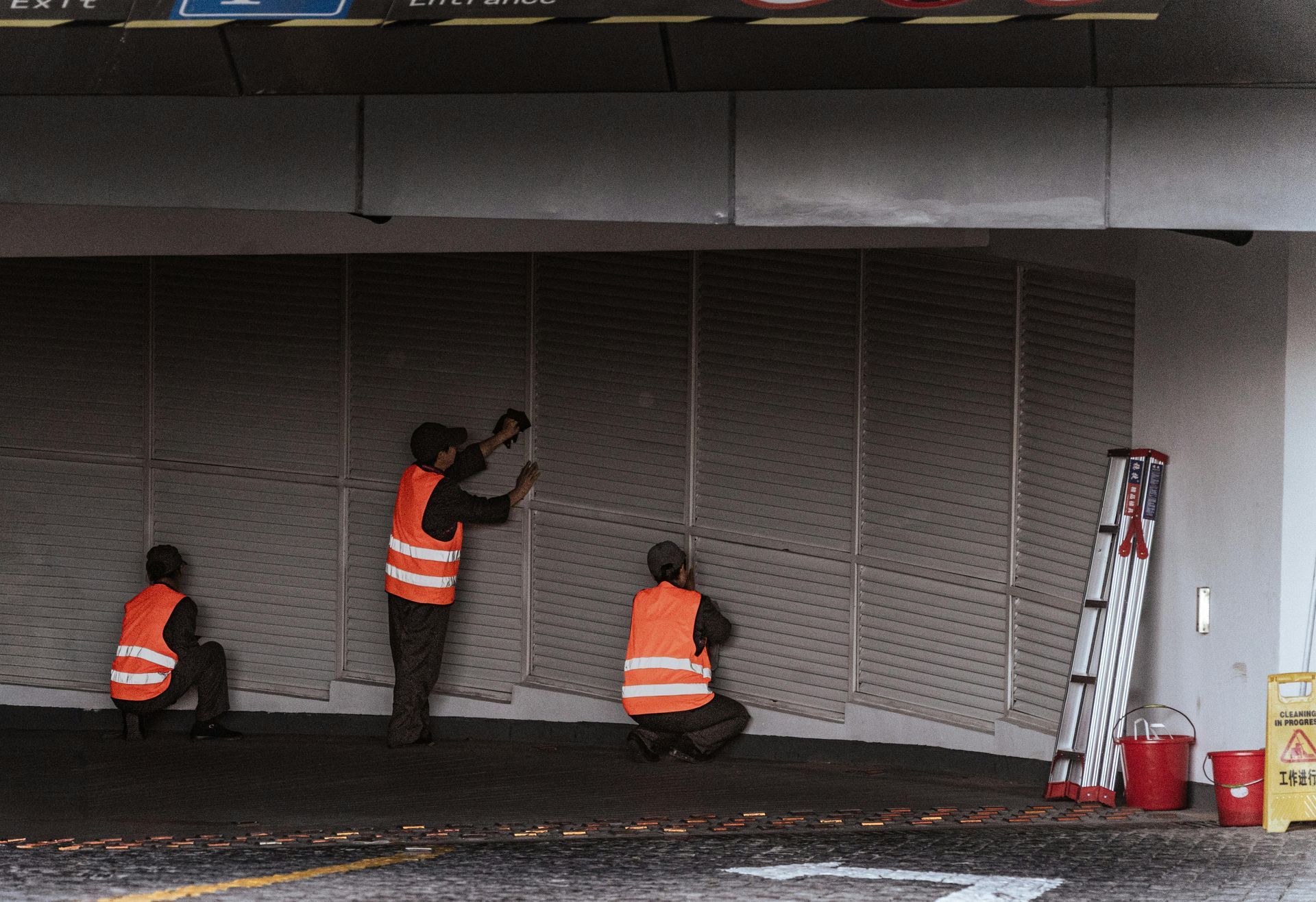 Three workers in orange vests install panels under a building overhang.