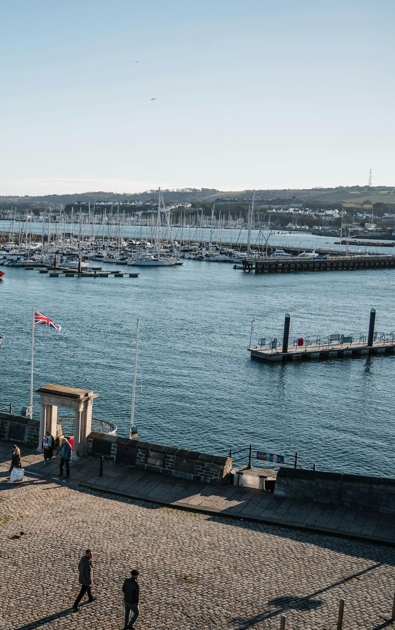 Harbor scene with boats, blue water, stone wall, and people walking on a cobblestone path.