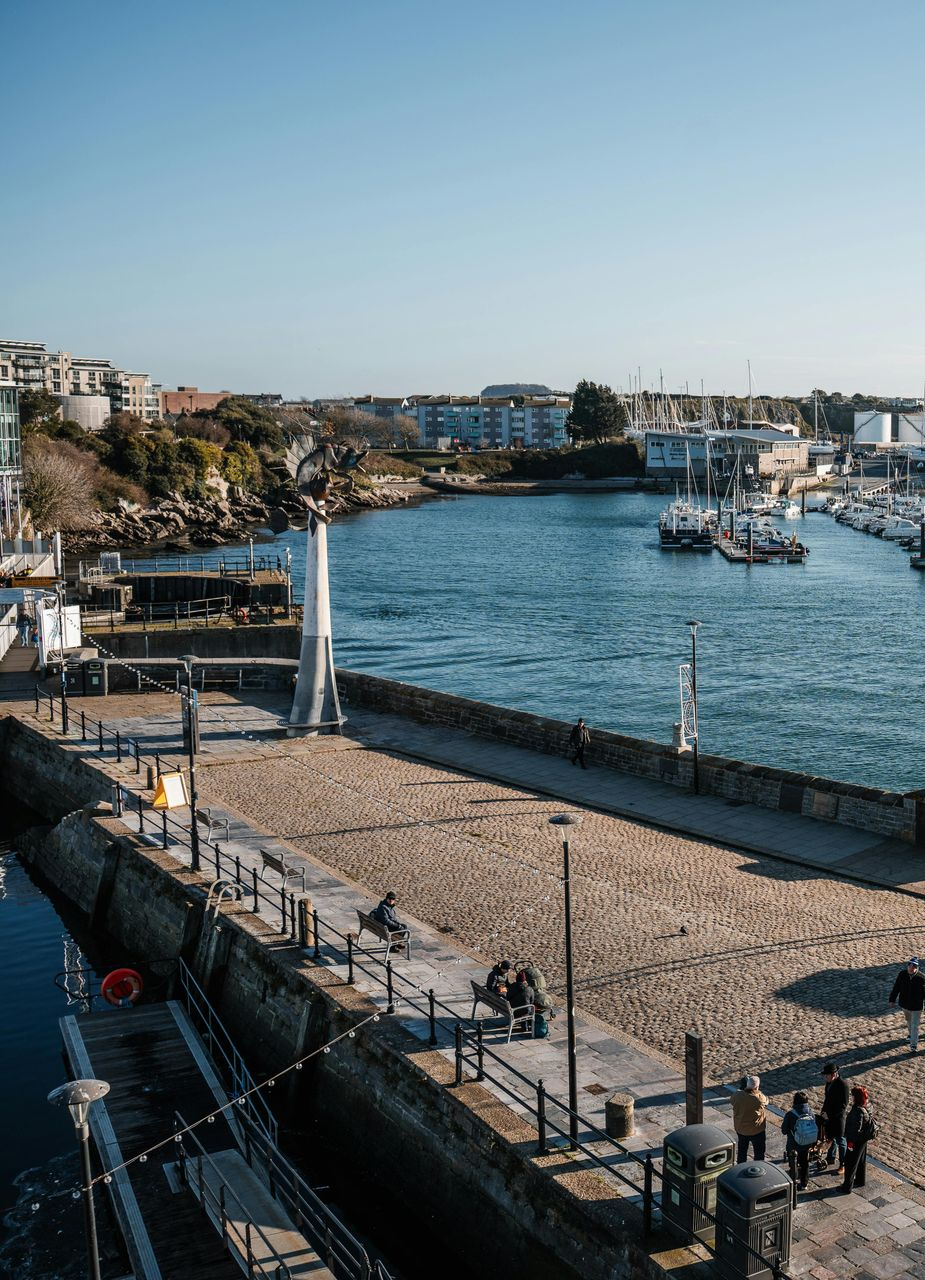 Harbor view: stone dock, boats, buildings, blue water, sunny day.