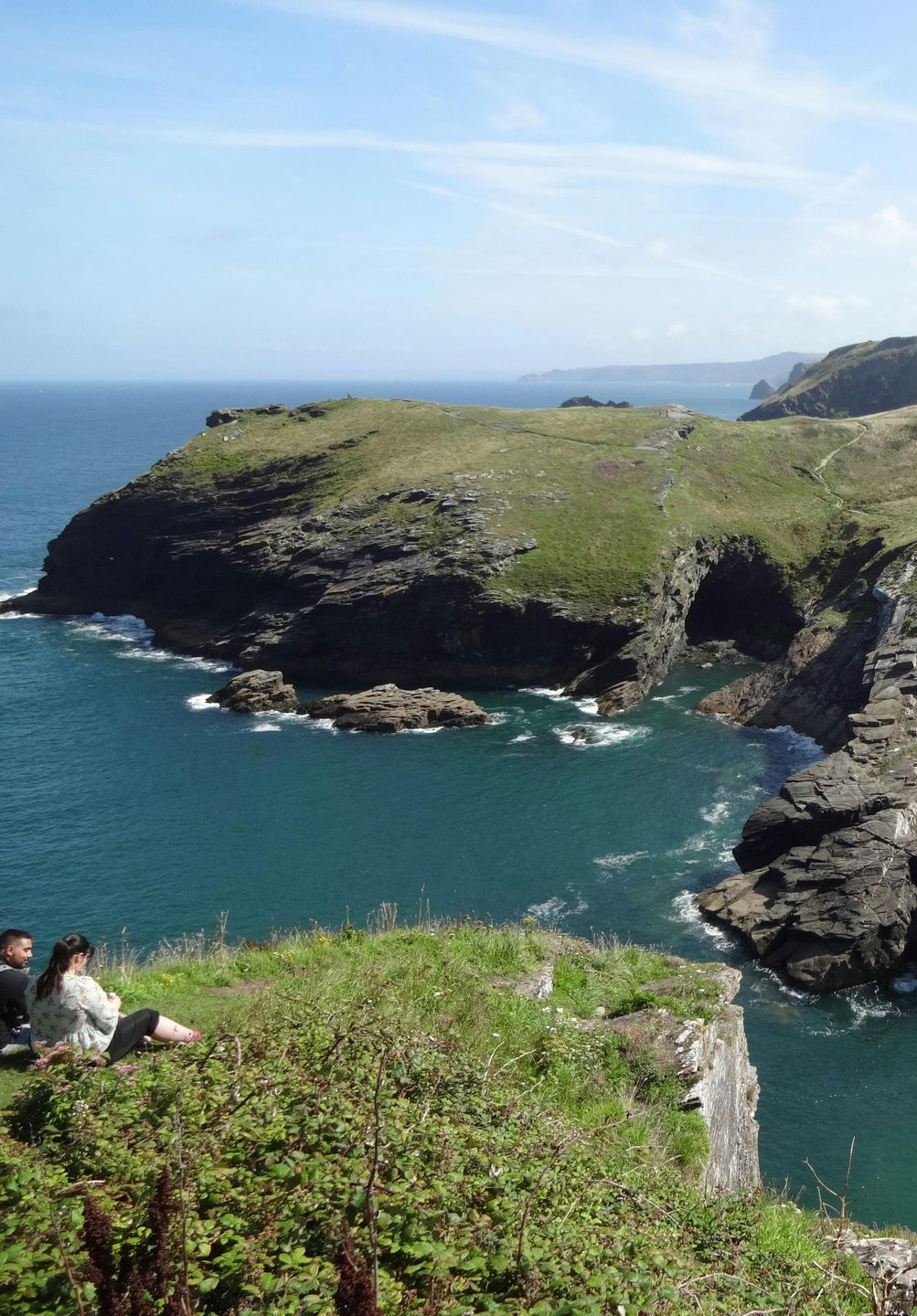 Coastal cliff with green grass, turquoise water, two people sitting. Sunny day.