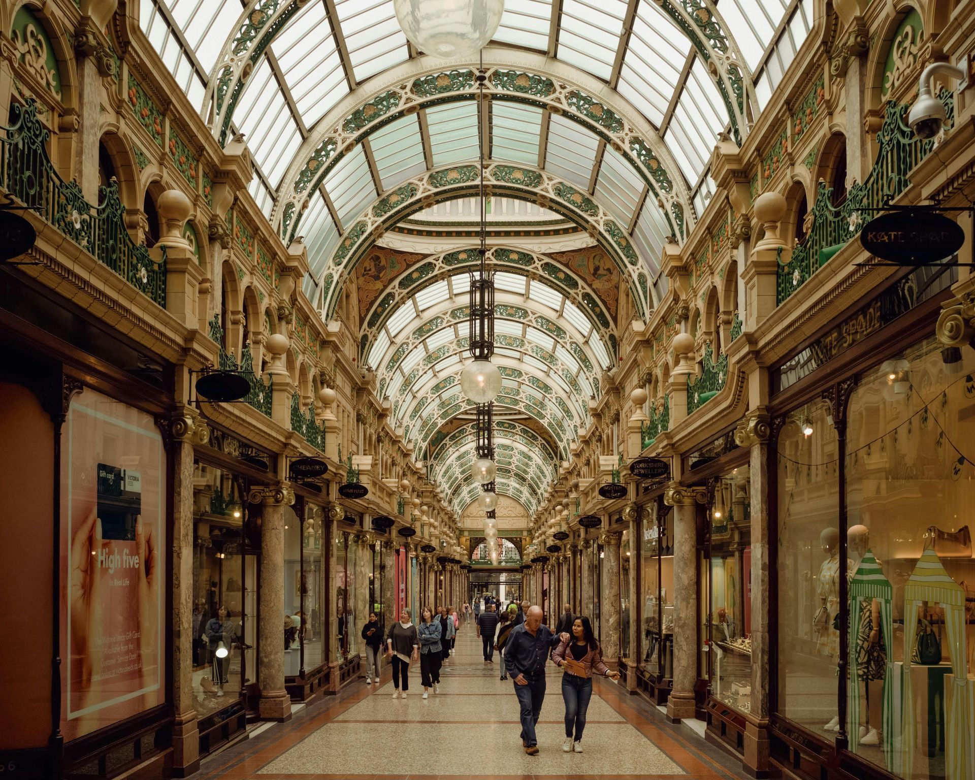 Arched glass-roofed shopping arcade with ornate facades, people walking.