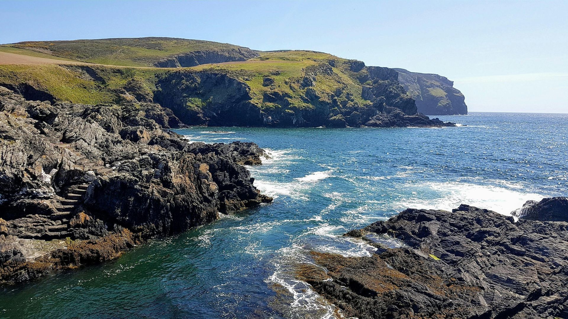 Rocky coastline meets the blue ocean under a sunny sky. Green grass tops a high cliff.