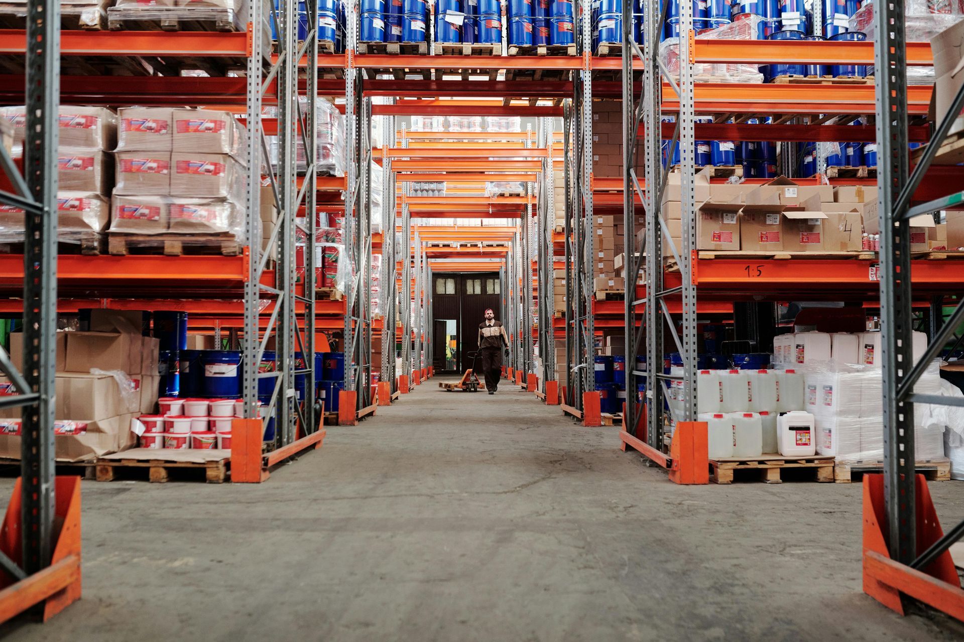 Warehouse interior with tall shelves stocked with various products; a person walks in the aisle.