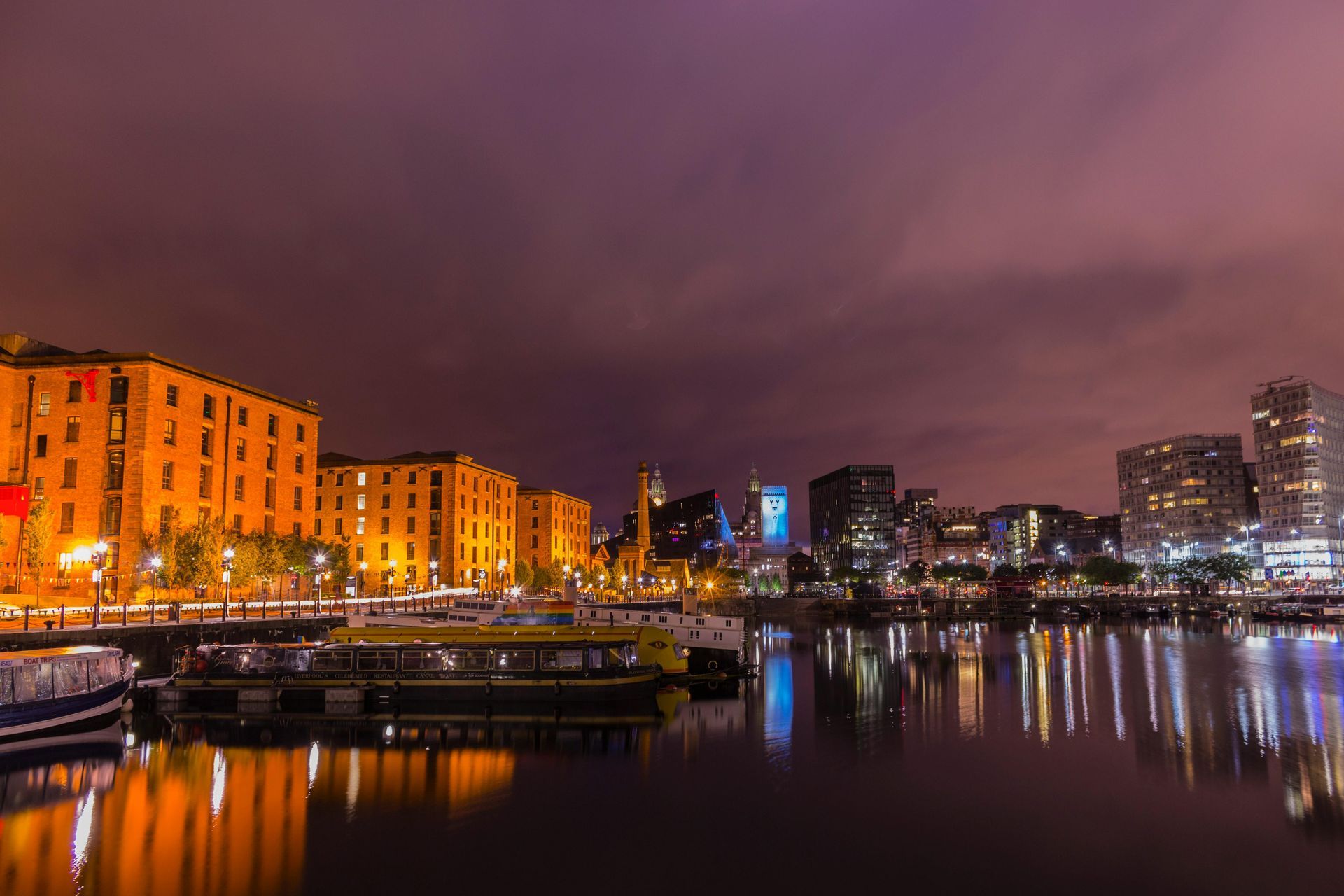 Night view of a harbor with illuminated buildings and reflections in the water. Dark purple sky.