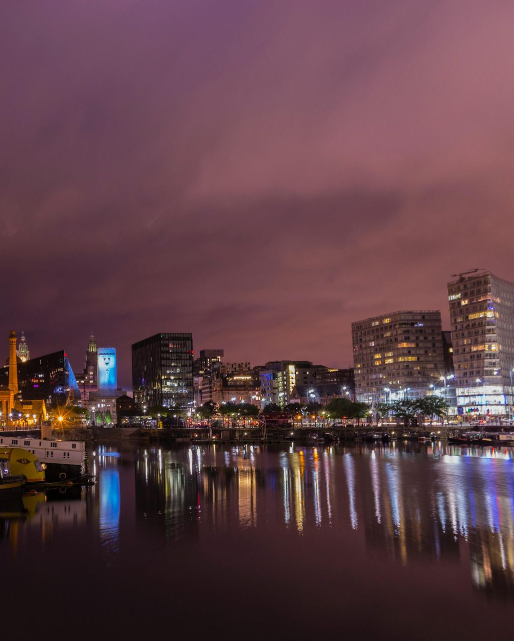 Nighttime cityscape with illuminated buildings reflecting in calm water under a purple-hued sky.
