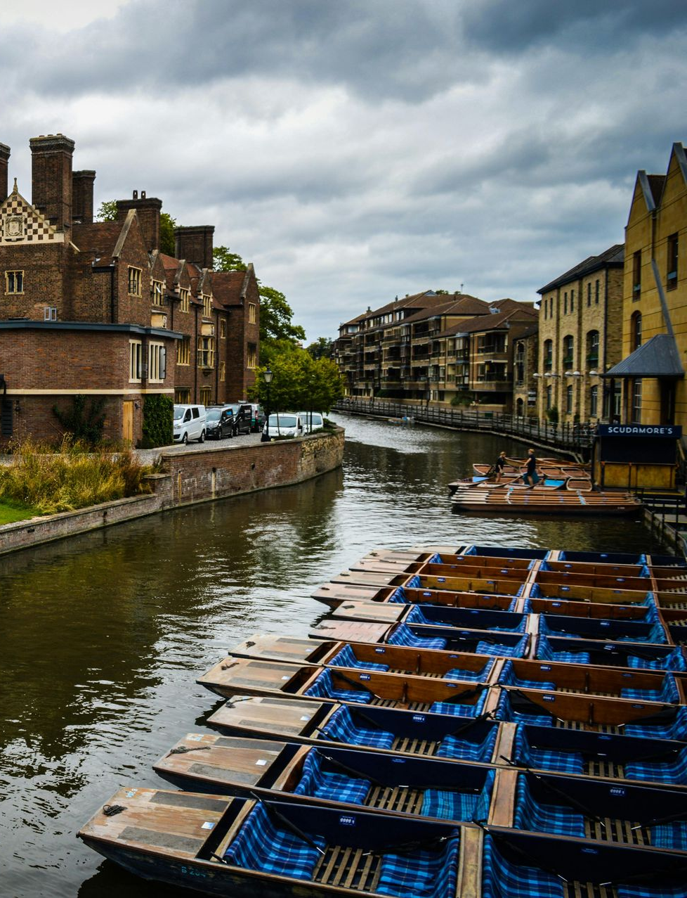 River Cam lined with punts, flowing past Cambridge buildings on a cloudy day.