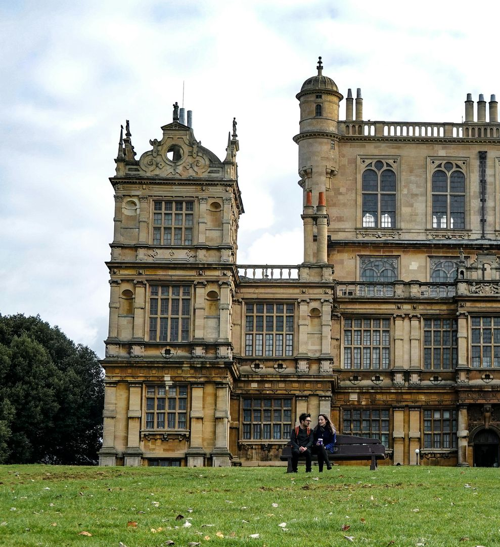 Two people sit on a bench in front of a large, ornate stone building, on a grassy hill.