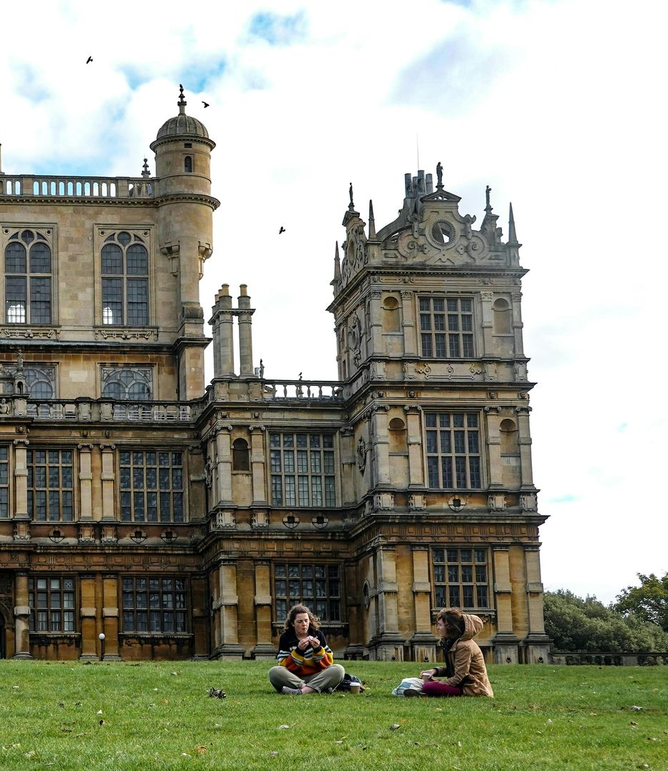 Two people sit on grass in front of a large, ornate building. Cloudy sky.