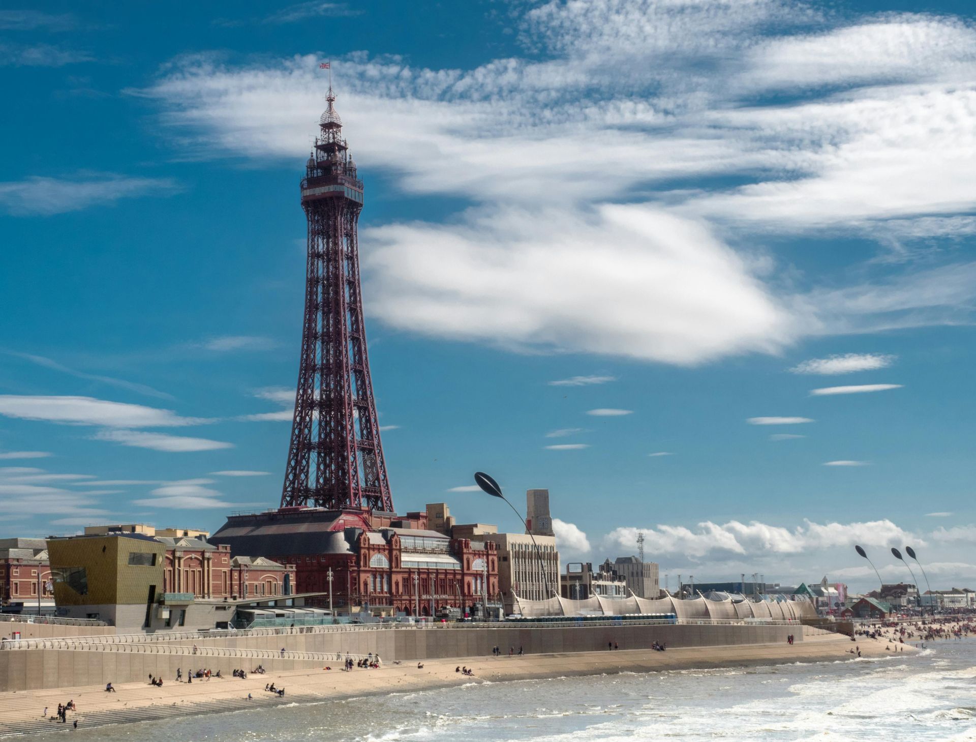 Blackpool Tower rises above buildings and beach on a sunny day with blue sky and some small planes flying.