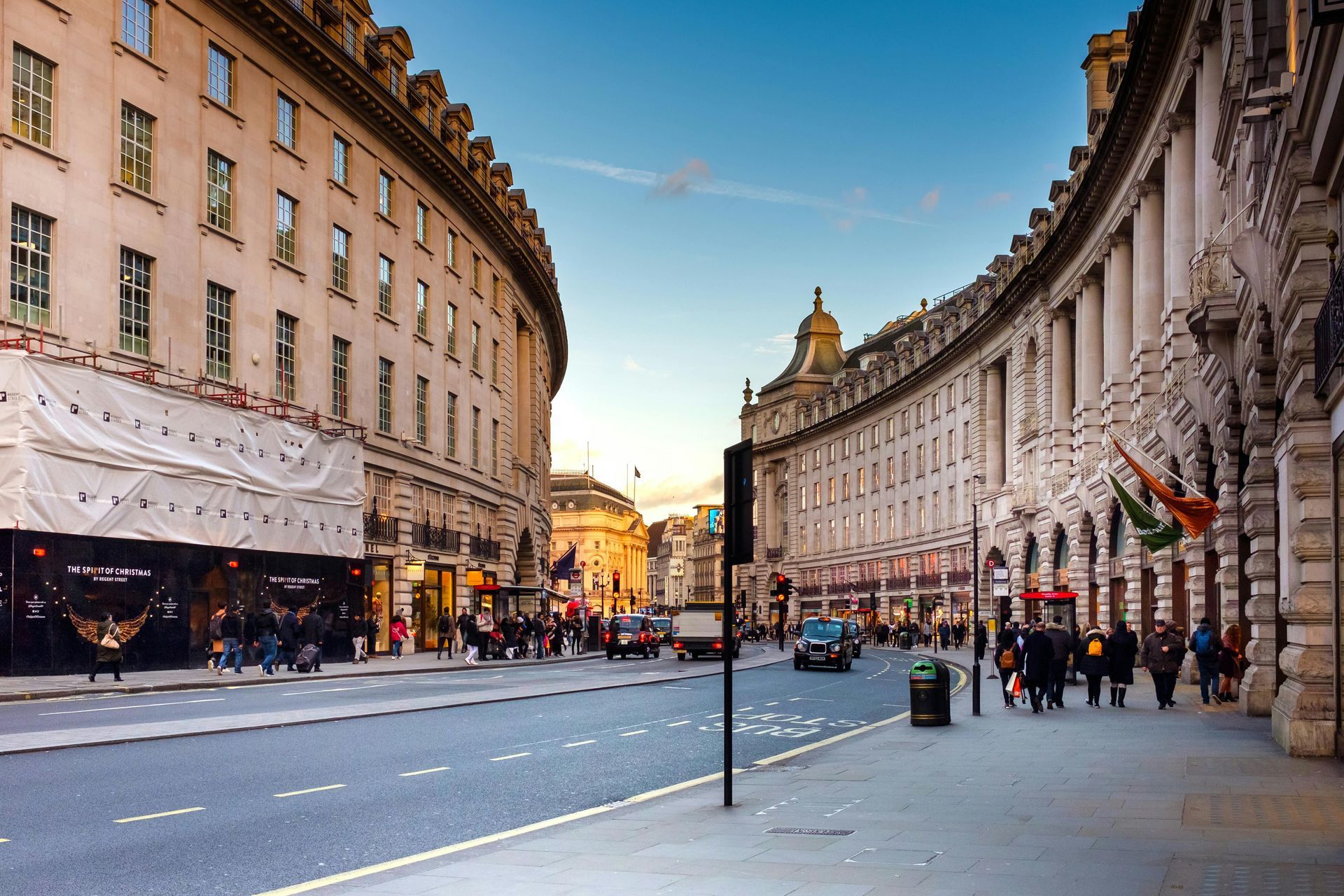 Curving street in London with buildings, shops, pedestrians, cars, and a blue sky.