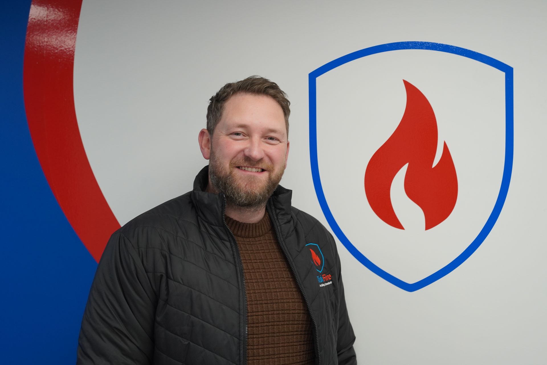 Man in black jacket and brown sweater smiling in front of a blue and white shield logo with a red flame.