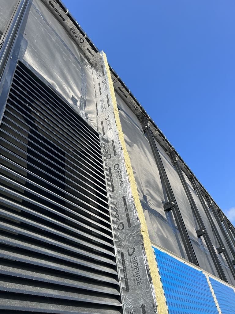 Exterior building facade with black horizontal louvers, insulation, and blue paneling against a blue sky.