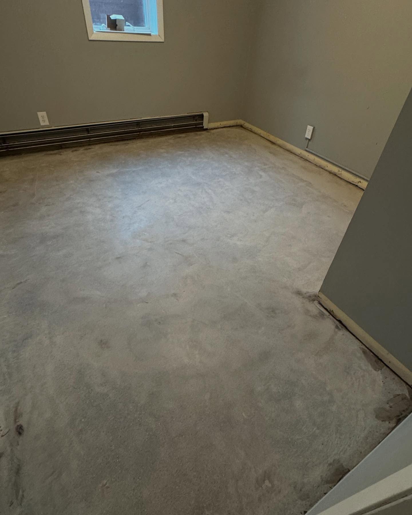 An empty room with gray walls and a concrete floor, featuring baseboard heating units and a small window.