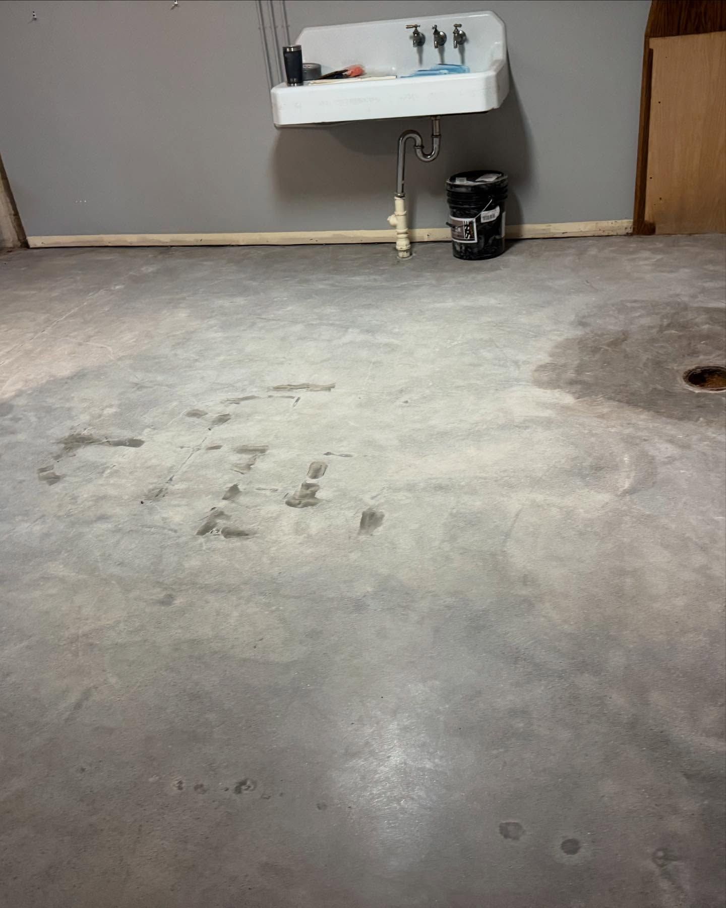 A concrete basement floor in front of a white utility sink mounted on a gray wall, with a small black bucket underneath.