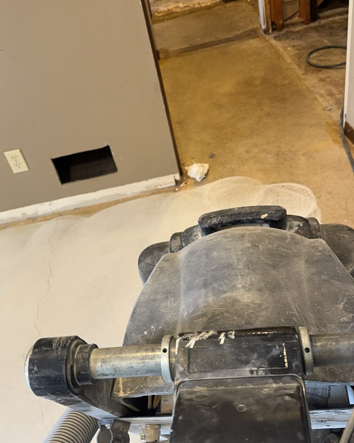 First-person view of a floor sander in use on a concrete basement floor near a gray wall.
