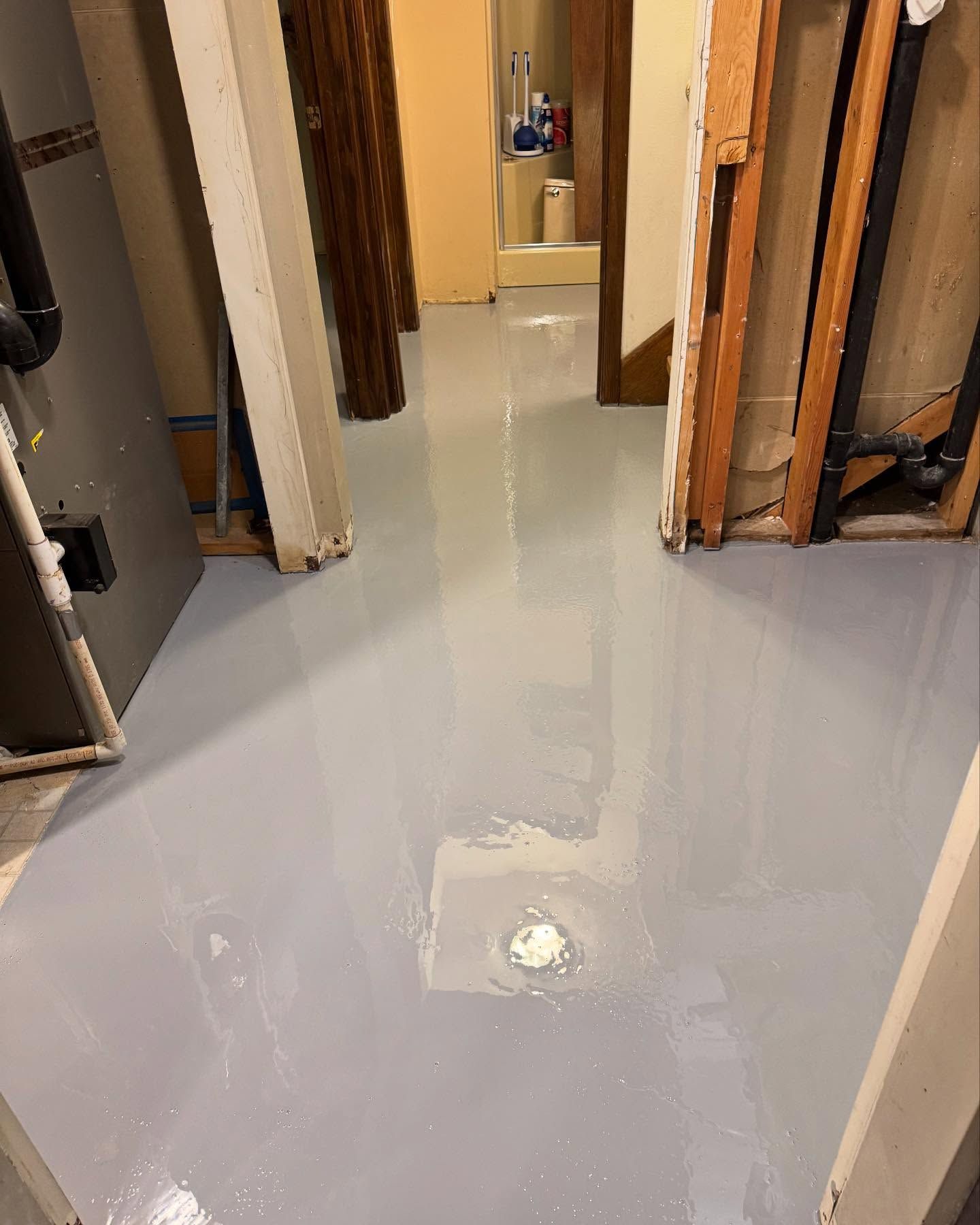 A freshly applied, glossy gray epoxy floor in an unfinished basement with a centered drain and wooden wall framing.