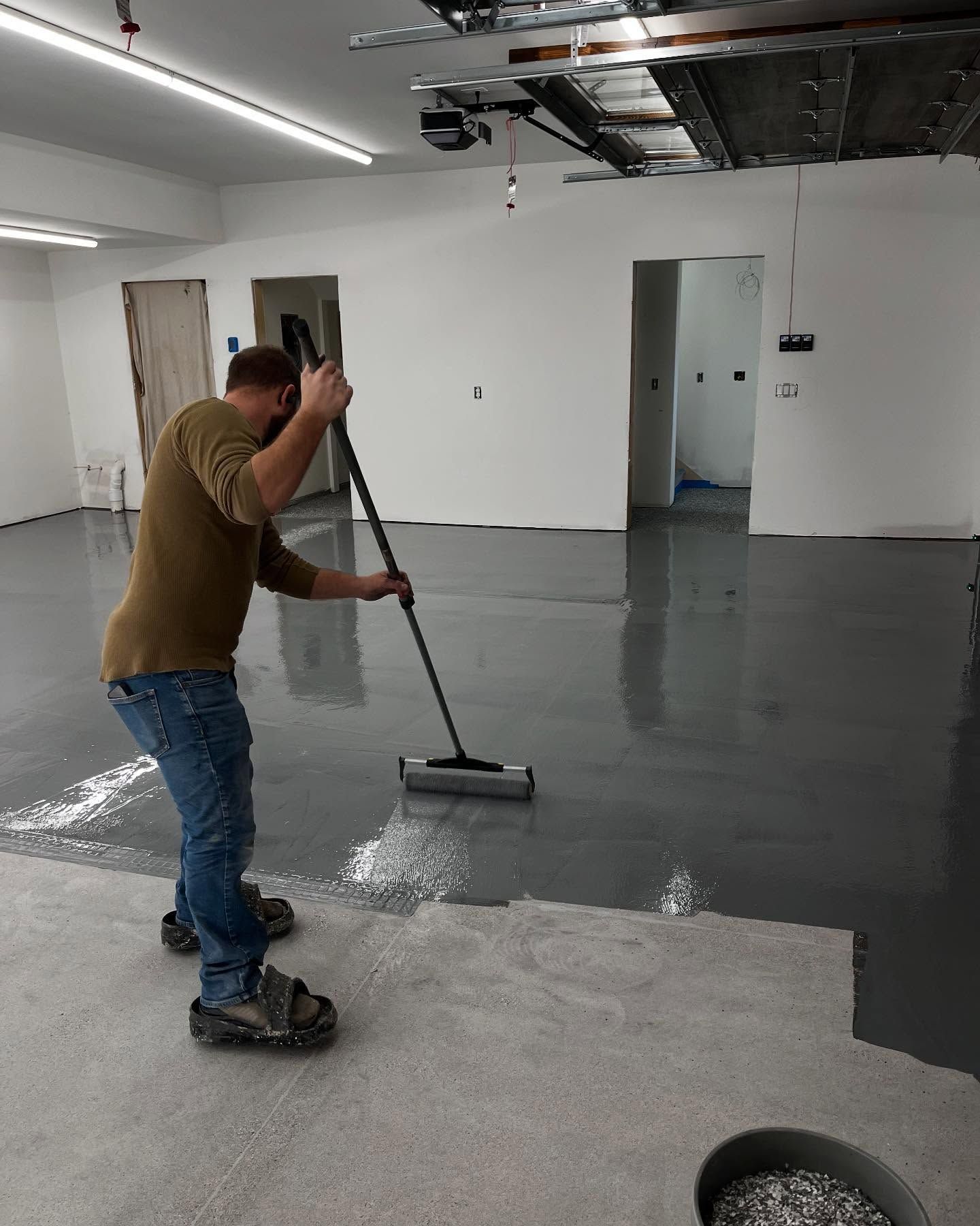 A person uses a long-handled roller to apply grey epoxy coating to a concrete floor in an unfinished room.